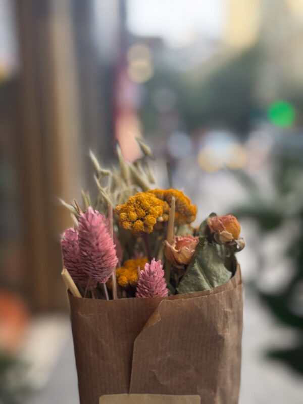 Απλότητα και κομψότητα στην πιο αγνή τους μορφή. Τα αποξηραμένα άνθη, ντυμένα με αποχρώσεις ώχρας, ροδακινί και dusty pink, αφηγούνται μια ιστορία φυσικής ομορφιάς. Δεν χρειάζονται φωνές· μόνο φως, σκιά και λίγο χαρτί craft για να ντύσουν τη διακριτική τους γοητεία. Ένα κομμάτι αγρού, μεταμορφωμένο σε λεπτομέρεια υψηλής αισθητικής.