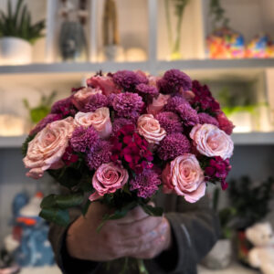 Στο επίκεντρο της σύνθεσης βρίσκονται τα ροζ ολλανδικά τριαντάφυλλα, γνωστά για τη φινέτσα και την πλούσια άνθισή τους. Με τα μεγάλα, μεταξένια πέταλα και τις απαλές διαβαθμίσεις του χρώματος, φέρνουν στο μπουκέτο μια αίσθηση πολυτέλειας και ρομαντικής νοσταλγίας. Δίπλα τους, τα χρυσάνθεμα σαντίνι προσθέτουν ένταση και δομή, με τα μικρά, σφαιρικά τους άνθη να δημιουργούν όμορφη υφή και οπτικό ενδιαφέρον. Ο συνδυασμός αυτός ολοκληρώνεται με το γαλλικό γαρύφαλλο, ένα άνθος με διαχρονική αξία στην ανθοδετική, που ενισχύει τον όγκο και το χρώμα, προσδίδοντας έναν τόνο κλασικής κομψότητας. Κάθε λουλούδι παίζει τον ρόλο του στη σύνθεση, δημιουργώντας ένα αποτέλεσμα αρμονικό και γεμάτο ζωή.