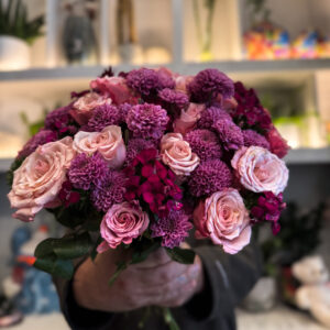 Στο επίκεντρο της σύνθεσης βρίσκονται τα ροζ ολλανδικά τριαντάφυλλα, γνωστά για τη φινέτσα και την πλούσια άνθισή τους. Με τα μεγάλα, μεταξένια πέταλα και τις απαλές διαβαθμίσεις του χρώματος, φέρνουν στο μπουκέτο μια αίσθηση πολυτέλειας και ρομαντικής νοσταλγίας. Δίπλα τους, τα χρυσάνθεμα σαντίνι προσθέτουν ένταση και δομή, με τα μικρά, σφαιρικά τους άνθη να δημιουργούν όμορφη υφή και οπτικό ενδιαφέρον. Ο συνδυασμός αυτός ολοκληρώνεται με το γαλλικό γαρύφαλλο, ένα άνθος με διαχρονική αξία στην ανθοδετική, που ενισχύει τον όγκο και το χρώμα, προσδίδοντας έναν τόνο κλασικής κομψότητας. Κάθε λουλούδι παίζει τον ρόλο του στη σύνθεση, δημιουργώντας ένα αποτέλεσμα αρμονικό και γεμάτο ζωή.