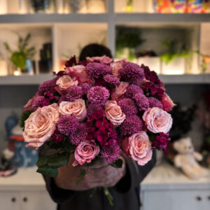 Στο επίκεντρο της σύνθεσης βρίσκονται τα ροζ ολλανδικά τριαντάφυλλα, γνωστά για τη φινέτσα και την πλούσια άνθισή τους. Με τα μεγάλα, μεταξένια πέταλα και τις απαλές διαβαθμίσεις του χρώματος, φέρνουν στο μπουκέτο μια αίσθηση πολυτέλειας και ρομαντικής νοσταλγίας. Δίπλα τους, τα χρυσάνθεμα σαντίνι προσθέτουν ένταση και δομή, με τα μικρά, σφαιρικά τους άνθη να δημιουργούν όμορφη υφή και οπτικό ενδιαφέρον. Ο συνδυασμός αυτός ολοκληρώνεται με το γαλλικό γαρύφαλλο, ένα άνθος με διαχρονική αξία στην ανθοδετική, που ενισχύει τον όγκο και το χρώμα, προσδίδοντας έναν τόνο κλασικής κομψότητας. Κάθε λουλούδι παίζει τον ρόλο του στη σύνθεση, δημιουργώντας ένα αποτέλεσμα αρμονικό και γεμάτο ζωή.