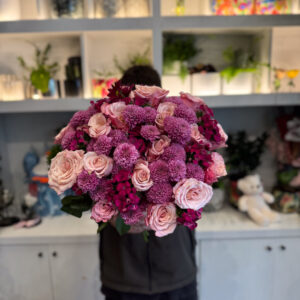 Στο επίκεντρο της σύνθεσης βρίσκονται τα ροζ ολλανδικά τριαντάφυλλα, γνωστά για τη φινέτσα και την πλούσια άνθισή τους. Με τα μεγάλα, μεταξένια πέταλα και τις απαλές διαβαθμίσεις του χρώματος, φέρνουν στο μπουκέτο μια αίσθηση πολυτέλειας και ρομαντικής νοσταλγίας. Δίπλα τους, τα χρυσάνθεμα σαντίνι προσθέτουν ένταση και δομή, με τα μικρά, σφαιρικά τους άνθη να δημιουργούν όμορφη υφή και οπτικό ενδιαφέρον. Ο συνδυασμός αυτός ολοκληρώνεται με το γαλλικό γαρύφαλλο, ένα άνθος με διαχρονική αξία στην ανθοδετική, που ενισχύει τον όγκο και το χρώμα, προσδίδοντας έναν τόνο κλασικής κομψότητας. Κάθε λουλούδι παίζει τον ρόλο του στη σύνθεση, δημιουργώντας ένα αποτέλεσμα αρμονικό και γεμάτο ζωή.