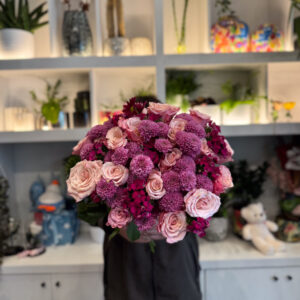 Στο επίκεντρο της σύνθεσης βρίσκονται τα ροζ ολλανδικά τριαντάφυλλα, γνωστά για τη φινέτσα και την πλούσια άνθισή τους. Με τα μεγάλα, μεταξένια πέταλα και τις απαλές διαβαθμίσεις του χρώματος, φέρνουν στο μπουκέτο μια αίσθηση πολυτέλειας και ρομαντικής νοσταλγίας. Δίπλα τους, τα χρυσάνθεμα σαντίνι προσθέτουν ένταση και δομή, με τα μικρά, σφαιρικά τους άνθη να δημιουργούν όμορφη υφή και οπτικό ενδιαφέρον. Ο συνδυασμός αυτός ολοκληρώνεται με το γαλλικό γαρύφαλλο, ένα άνθος με διαχρονική αξία στην ανθοδετική, που ενισχύει τον όγκο και το χρώμα, προσδίδοντας έναν τόνο κλασικής κομψότητας. Κάθε λουλούδι παίζει τον ρόλο του στη σύνθεση, δημιουργώντας ένα αποτέλεσμα αρμονικό και γεμάτο ζωή.