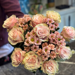 Στο επίκεντρο της σύνθεσης βρίσκονται τα τριαντάφυλλα Εκουαδόρ — ίσως τα πιο εκλεκτά τριαντάφυλλα στον κόσμο, γνωστά για τα μεγάλα τους άνθη, τα σταθερά τους στελέχη και τα πυκνά, μεταξένια πέταλα. Οι απαλές σομόν αποχρώσεις τους διαβαθμίζονται από ένα απαλό ροζ στο κέντρο μέχρι ένα ελαφρύ κρεμ στις άκρες, δημιουργώντας μια φυσική μετάβαση που αποπνέει τρυφερότητα και φως.