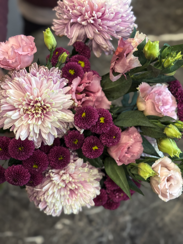 Οι λυσίανθοι στις απαλές ροζ αποχρώσεις τους λειτουργούν σαν «γέφυρα» ανάμεσα στα μωβ και τα λιλά χρυσάνθεμα, ενοποιώντας τη χρωματική παλέτα με φυσική χάρη. Είναι λουλούδια με διάρκεια, που διατηρούν τη φρεσκάδα τους για πολλές ημέρες, κάτι που κάνει την ανθοδέσμη ιδανική επιλογή τόσο για δώρο όσο και για διακόσμηση.