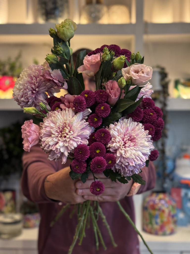 Τα σαντίνι χρυσάνθεμα — γνωστά για το στρογγυλό, μικρό τους άνθος — προσδίδουν όγκο και ένταση στη σύνθεση. Με το βαθύ, πλούσιο μωβ τους, δημιουργούν ένα δυνατό οπτικό σημείο, αναδεικνύοντας τα υπόλοιπα άνθη γύρω τους.