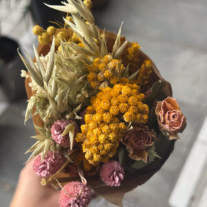 Μια μικρή συμφωνία από χρώμα και υφή — αποξηραμένα άνθη σε αποχρώσεις ροζ και χρυσού, σαν στιγμιότυπο από το φως του δειλινού. Οι θερμοί τόνοι του κίτρινου αγκαλιάζουν τα dusty pink πέταλα, ενώ το φυσικό πράσινο των στελεχών χαρίζει ισορροπία και κίνηση. Μια ανθοδέσμη που μοιάζει να αιχμαλωτίζει τη στιγμή που ο ήλιος χαμηλώνει και όλα γίνονται πιο απαλά, πιο αληθινά, πιο όμορφα.