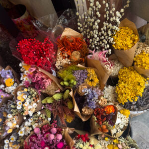 Ένα παζλ από αποξηραμένα άνθη — ρουμπινί, ώχρα, λιλά και απαλό ροζ — σχηματίζουν έναν πίνακα γεμάτο κίνηση και ζωή. Κάθε ανθοδέσμη είναι μια μικρή ιστορία: άλλες μιλούν με ένταση, άλλες ψιθυρίζουν απαλότητα. Μαζί, δημιουργούν μια χρωματική συμφωνία που μοιάζει περισσότερο με καμβά παρά με πάγκο ανθοπωλείου.