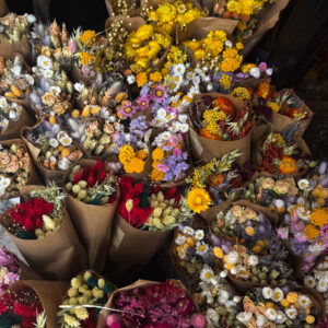 Στην άκρη του δρόμου, ένα μικρό θαύμα χρώματος. Πληθώρα από άνθη — αμάραντα, λυσίανθοι, λεβάντες και μαργαρίτες — σχηματίζουν μια αυθόρμητη σύνθεση με υψηλή αισθητική. Είναι η γοητεία της ατέλειας, εκεί όπου το οργανικό και το εκλεπτυσμένο συναντιούνται. Rustic κομψότητα με αύρα Παρισιού.