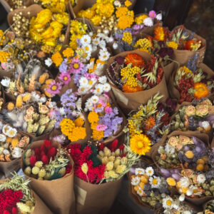 Κάποτε άνθισαν στο φως. Τώρα, φυλάνε μέσα τους τη μνήμη του καλοκαιριού. Οι χρυσοί, ροδακινί και μοβ τόνοι συνθέτουν ένα τοπίο γεμάτο ζεστασιά και ηρεμία. Τα αποξηραμένα άνθη δεν κουβαλούν φθορά, αλλά διάρκεια — μια ήσυχη, διαχρονική πολυτέλεια που ακουμπά την ψυχή.